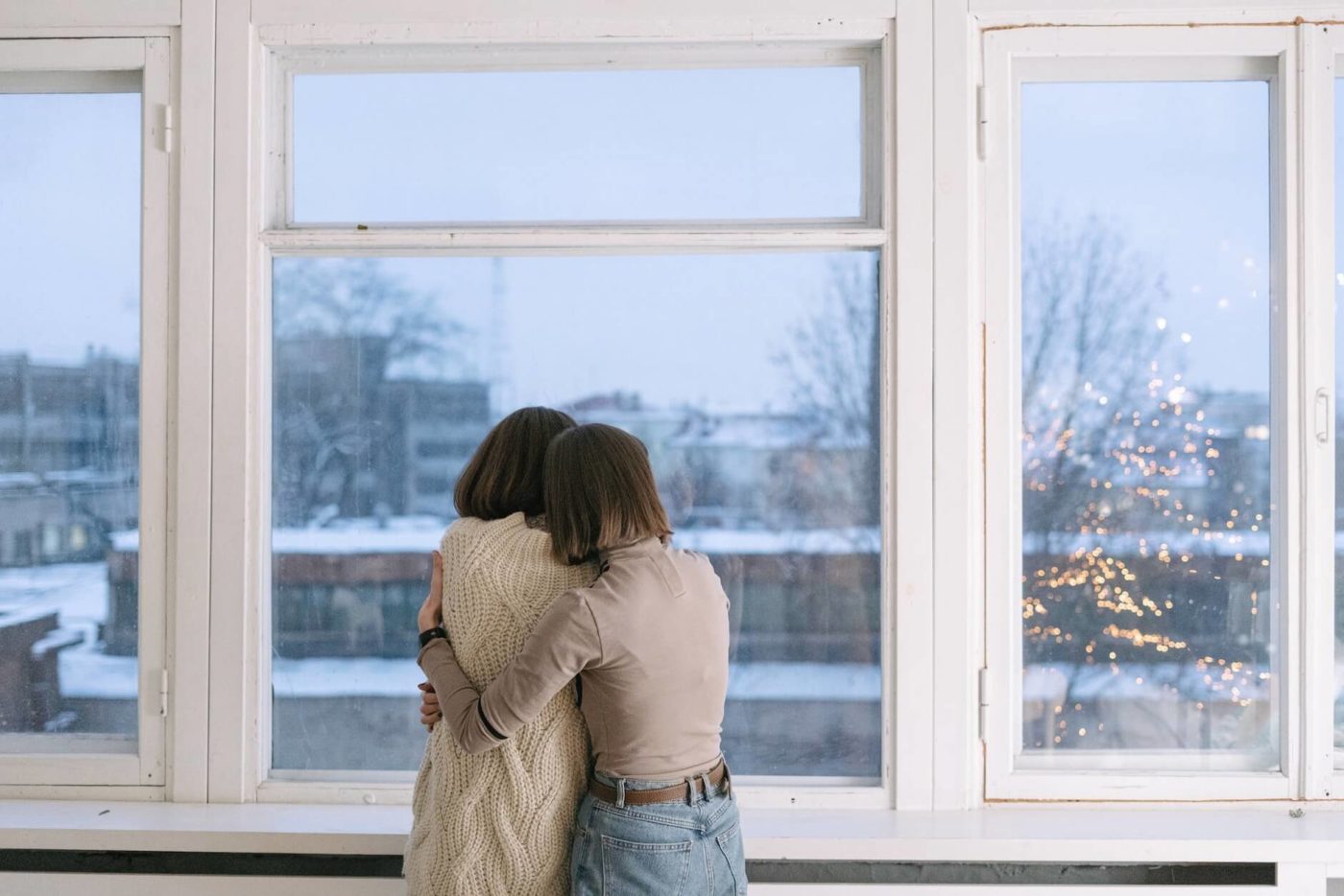 two women standing in front of a window and hugging while there's snow outside