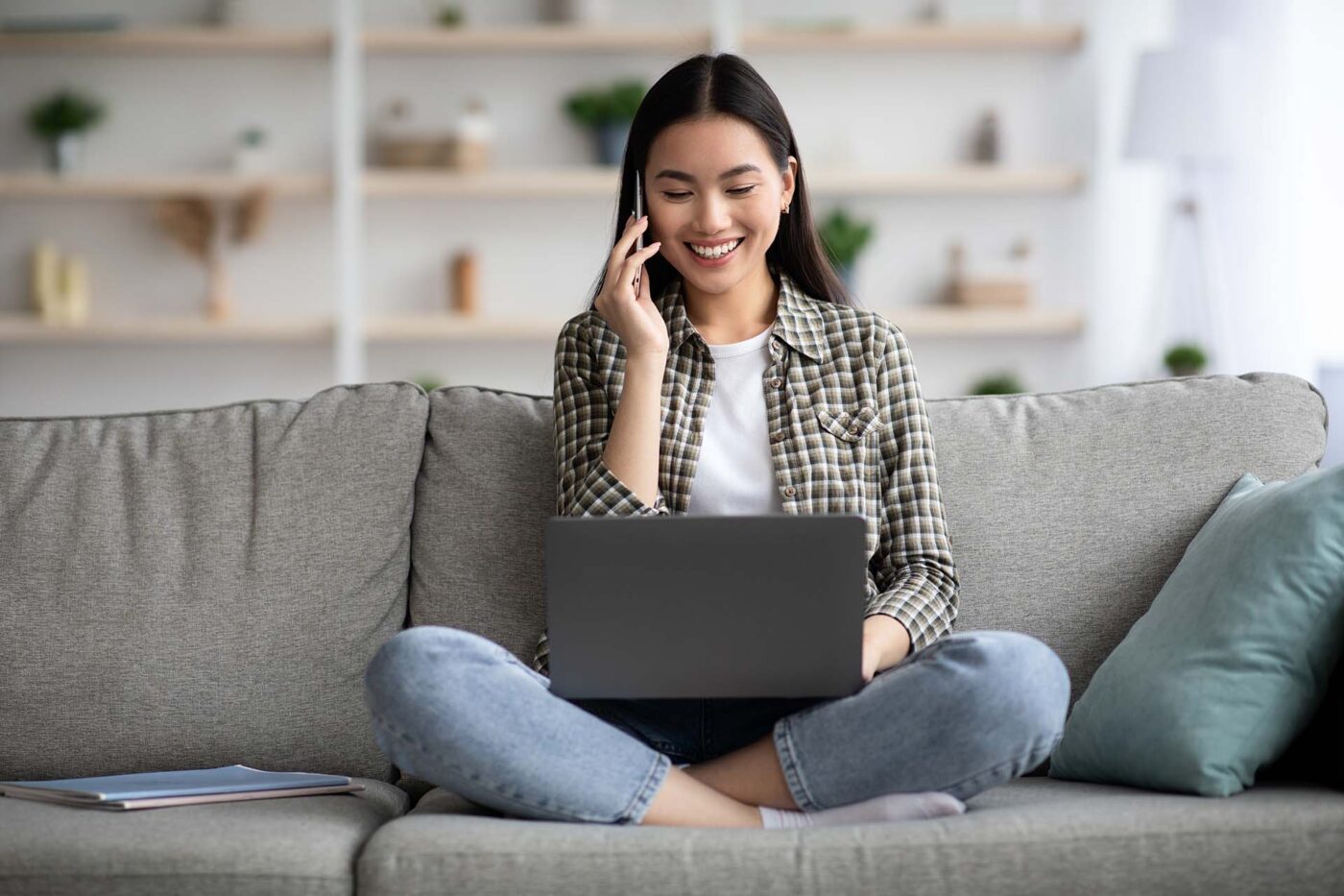 Relaxed asian woman with laptop looking for job opportunities online, having phone conversation with employer, happy young korean lady sitting on couch at home, using notebook, copy space